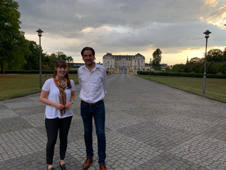 Ein Mann und eine Frau stehen lächelnd auf einem Kopfsteinpflasterweg vor einem großen, gelben Schloss bei Sonnenuntergang.