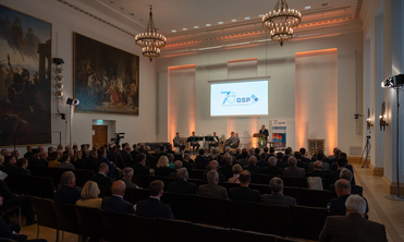 Publikum in einem Saal schaut einem Redner am Pult auf einer Bühne zu, hinter ihm eine Leinwand mit „70 Jahre GSP“ und Musiker.