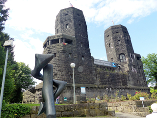 Die zwei dunklen Steintürme des Friedensmuseums Brücke von Remagen, mit abstrakter Skulptur im Vordergrund und Flaggen.