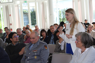 Junge Frau spricht auf Konferenz in ein Mikrofon, vor einem gemischten Publikum, darunter ein Mann in Militäruniform.