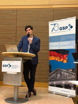 Ein Mann hält einen Vortrag an einem Rednerpult vor einem Banner der Gesellschaft für Sicherheitspolitik (GSP) mit deutscher Flagge und Reichstag.