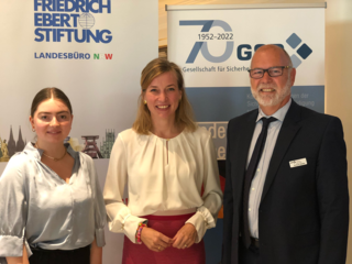 Drei lächelnde Personen, zwei Frauen und ein Mann, vor Bannern der Friedrich-Ebert-Stiftung und der GSP.
