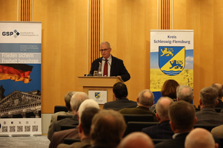 Ein Redner spricht vor einem Publikum, flankiert von Bannern der Gesellschaft für Sicherheitspolitik und Kreis Schleswig-Flensburg.