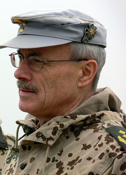 Mann in deutscher Militäruniform, grauer Mütze mit Edelweiß-Abzeichen, Flecktarn-Jacke, Brille und Schnurrbart.