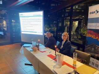 Eine Frau und ein Mann sitzen an einem Tisch bei einer GSP-Veranstaltung mit Projektion und Banner. Die Frau lächelt.