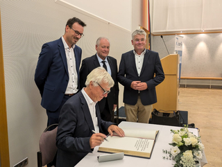 Ein Mann mit weißem Haar unterschreibt ein großes Buch, beobachtet von drei Männern in Anzügen.