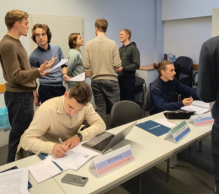 Studenten bei einer Model-UN-Simulation. Einige stehen und diskutieren, andere sitzen mit Länderschildern (Russland, Sierra Leone, Slowenien).