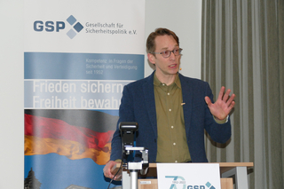 Ein Mann mit Brille und blauer Jacke spricht an einem Pult vor einem GSP-Banner mit deutscher Flagge.