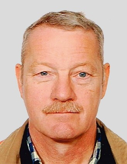 Kopfaufnahme eines älteren Mannes mit grauem Haar, blauen Augen und Schnurrbart, der ein kariertes Hemd und eine braune Jacke trägt.