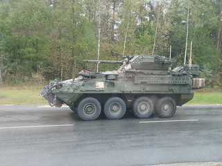 Ein grünes gepanzertes Stryker Mobile Gun System mit großer Kanone fährt auf nasser Straße neben einem Wald.
