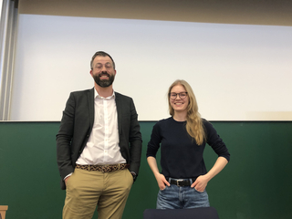 Lächelnder bärtiger Mann mit Brille und Frau mit langen blonden Haaren und Brille in einem Klassenzimmer vor Tafel.