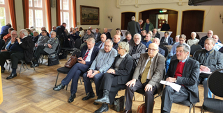 Ältere Männer in einem großen Raum mit Holzboden und Fenstern, einige in Uniform, sitzen und hören einer Veranstaltung zu.
