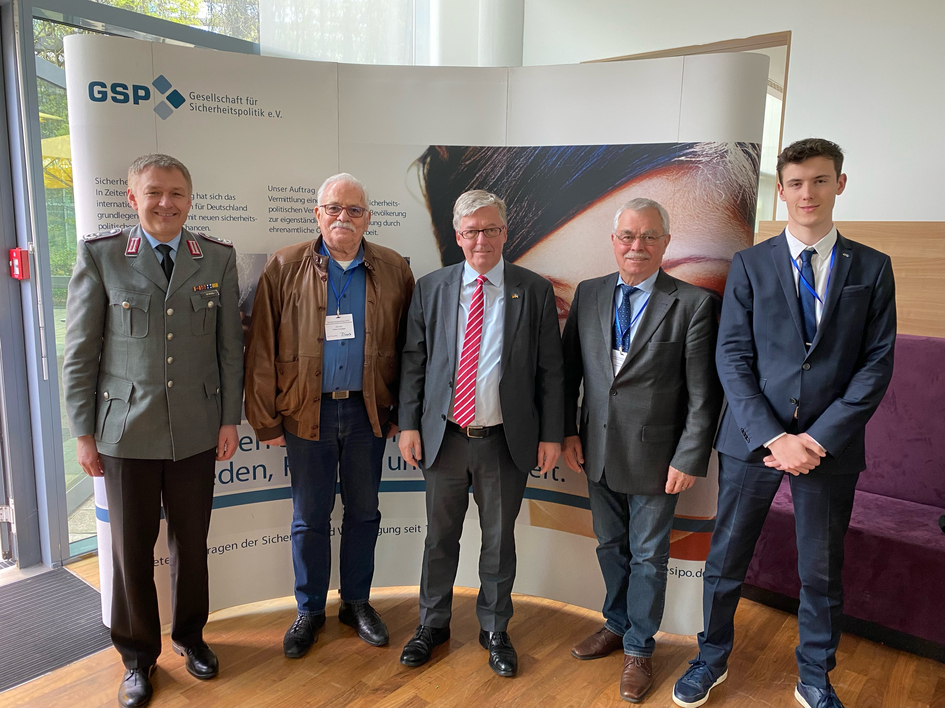 Fünf Männer verschiedener Generationen und Berufe, darunter ein Soldat in Uniform und ein junger Mann im Anzug, stehen vor einem GSP-Banner.