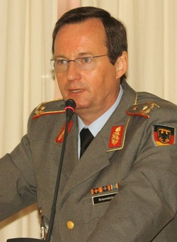 Generalmajor Jörg Braunstein der Bundeswehr spricht in olivgrüner Uniform mit Rangabzeichen und Deutschlandflagge ins Mikrofon.