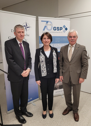 Drei Personen in Geschäftskleidung vor GSP-Bannern, die 70 Jahre Sicherheitspolitik feiern.