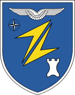 Wappen auf blauem Schild mit weißen Flügeln, NATO-Stern, gelben Blitzen und weißem Burgturm.