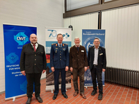 Vier Männer, einer in Uniform, lächeln vor Bannern der DWT, Gesellschaft für Sicherheitspolitik und des BundeswehrVerbandes.