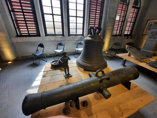 Eine alte Kanone, eine große Glocke und eine Statue auf Holzpodesten in einem historischen Raum mit hohen Fenstern.