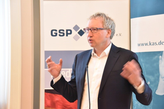 Ein Mann mit Brille und hellem Haar spricht und gestikuliert mit den Händen vor Bannern von GSP und www.kas.de.