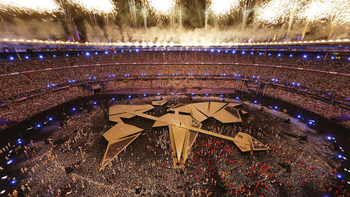 Feuerwerk über einem gefüllten Olympiastadion mit leuchtenden Ringen, geometrischer Bühne und Darstellern auf dem Feld.