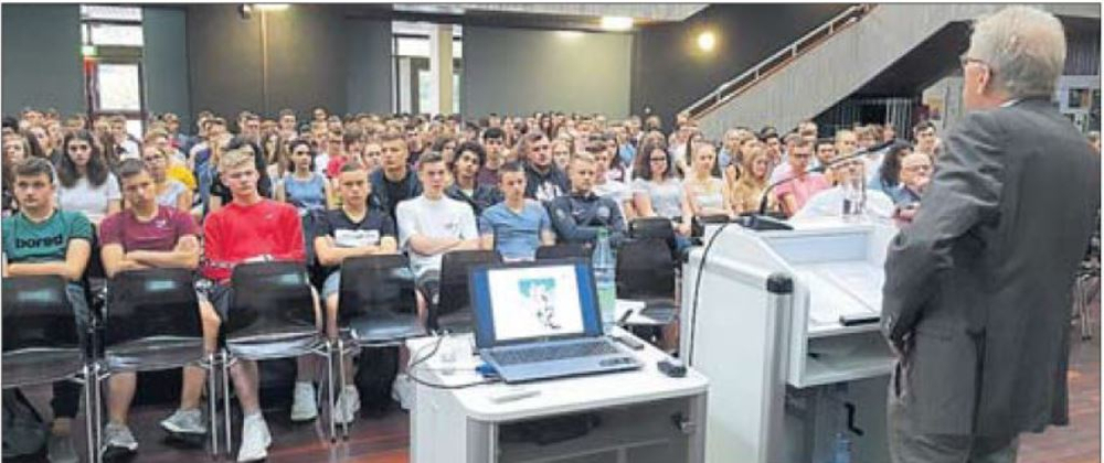 Ein Dozent hält einen Vortrag vor einer großen Gruppe junger Studierender in einem Hörsaal; ein Laptop zeigt eine Präsentation.