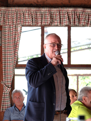 Ein Mann im dunklen Sakko mit Brille spricht in ein Mikrofon. Im Hintergrund sind ein Publikum und karierte Vorhänge.