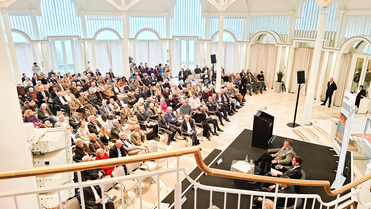 Überblick über eine Konferenzhalle mit großem Publikum und Rednern auf einer Bühne, viele Fenster im hellen Raum.