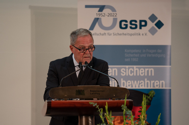 Ein Mann mit Brille spricht am Rednerpult des Bayerischen Landtags vor einem Banner der GSP zum 70-jährigen Jubiläum.