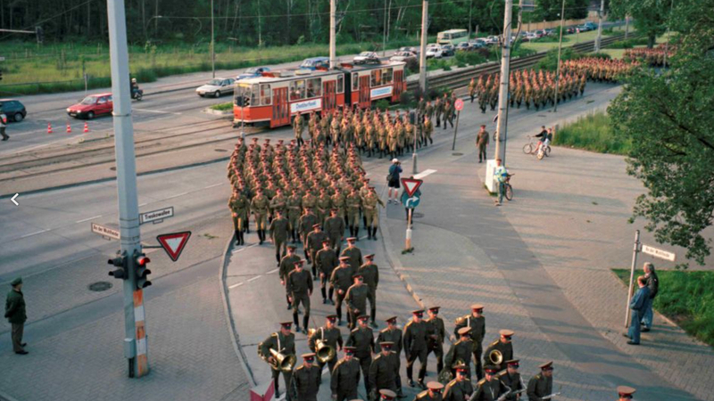 Militärparade auf der Treskowallee mit marschierenden Soldaten, einer roten Straßenbahn und Autos.