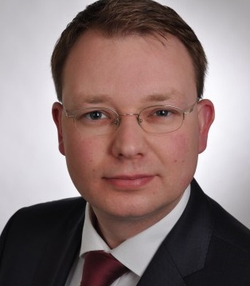 Kopfaufnahme eines Mannes mit Brille, kurzem braunem Haar, dunklem Anzug und roter Krawatte, der in die Kamera blickt.
