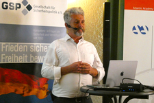 Mann mit Headset spricht vor GSP-Banner und deutscher Flagge bei einer Sicherheitspolitik-Veranstaltung. Laptop auf Tisch.