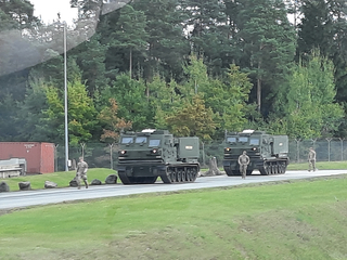 Zwei militärische MLRS-Fahrzeuge und Soldaten auf einer Straße neben einem Wald, mit einem roten Container und Zaun im Hintergrund.