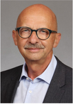 Porträt eines glatzköpfigen Mannes mit dunkler Brille, Schnurrbart, hellblauem Hemd und dunklem Sakko.