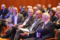 Eine Gruppe von Teilnehmern, hauptsächlich Männer in Anzügen, sitzt aufmerksam in Reihen bei einer formellen Konferenz oder Besprechung.