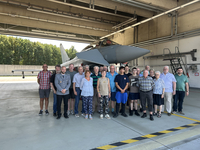 Gruppe vielfältiger Personen posiert vor einem grauen Eurofighter Typhoon Kampfjet in einem Hangar.