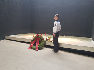 Ein Seemann in Uniform steht in einem feierlichen Gedenkraum vor einem Blumenkranz mit rotem Band und Kerzen.