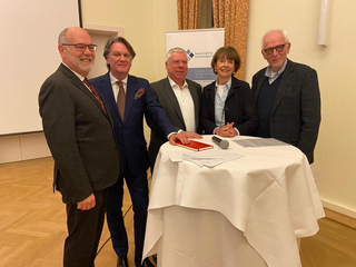 Fünf lächelnde Personen an einem Tisch, im Hintergrund ein Banner der Gesellschaft für Sicherheitspolitik.