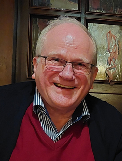 Ein glücklicher älterer Mann mit grauem Haar und Brille lächelt breit, trägt einen roten Pullover und ein gestreiftes Hemd.