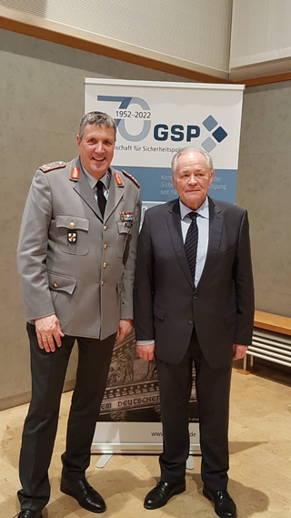 Zwei Männer, einer in Uniform, einer im Anzug, posieren vor einem GSP-Banner zum 70-jährigen Jubiläum der Sicherheitspolitik.