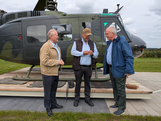 Drei Männer unterhalten sich lächelnd neben einem dunkelgrünen Militärhubschrauber auf einer Holzplattform im Freien.