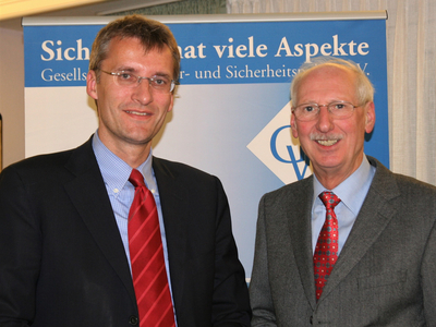 Zwei lächelnde Männer in Anzügen und Krawatten vor einem blauen Banner mit der Aufschrift 'Sicherheit hat viele Aspekte'.