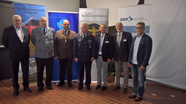 Sieben Männer, teils in Uniform, Vertreter deutscher Verteidigungs- und Sicherheitsorganisationen, posieren vor Bannern.
