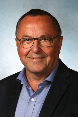 Porträt eines lächelnden älteren Mannes mit kurzen Haaren, Brille, blauem Hemd und dunklem Sakko.
