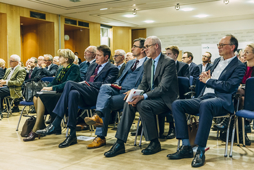Teilnehmer in Geschäftskleidung, hauptsächlich Männer, sitzen im Publikum bei einer formellen Konferenz oder Veranstaltung.