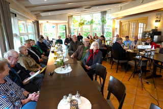 Eine große Gruppe von Menschen, hauptsächlich Senioren, versammelt in einem hellen Restaurantraum mit langen Tischen und großen Fenstern.