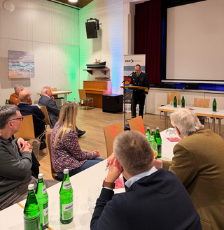 Mann in Uniform hält Vortrag vor Publikum bei einer GSP-Veranstaltung, mit grüner Beleuchtung und Schiffsmodell.