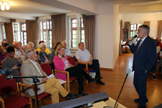 Ein Mann im Anzug spricht mit Mikrofon vor einem sitzenden Publikum in einem hellen Raum mit Fenstern.