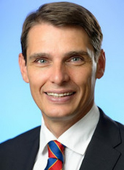 Kopfaufnahme eines lächelnden Mannes mit kurzen braunen Haaren, blauen Augen, in dunklem Anzug und gestreifter Krawatte vor blauem Hintergrund.