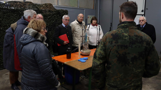 Ein Soldat erklärt einer Gruppe Zivilisten Laborgeräte an einem Tisch in einer Halle mit Tarnnetzen.