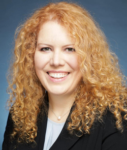 Porträt einer lächelnden Frau mit lockigem rotblondem Haar, schwarzem Blazer und Perlenkette vor blauem Hintergrund.
